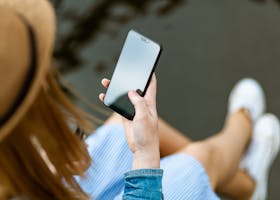A young woman holding a smartphone while sitting outdoors in summer, capturing a moment of relaxation and technology use.
