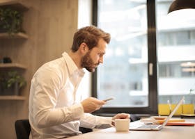 Focused businessman working on laptop while checking smartphone in modern office.